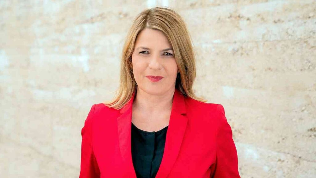 Frau mit blonden Haaren trägt einen roten Blazer und steht vor einer hellen Wand.