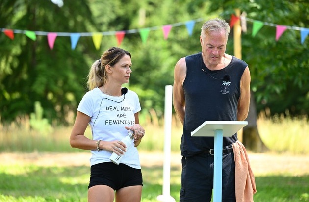 Frau mit T-Shirt 'Real Men Are Feminists' und Mann in ärmellosem Shirt stehen an einem Podium im Freien.