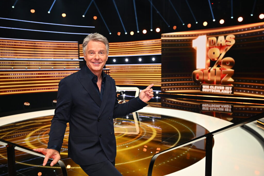 Moderator Jörg Pilawa präsentiert das 1% Quiz in einem modernen Fernsehstudio mit beleuchtetem Hintergrund.