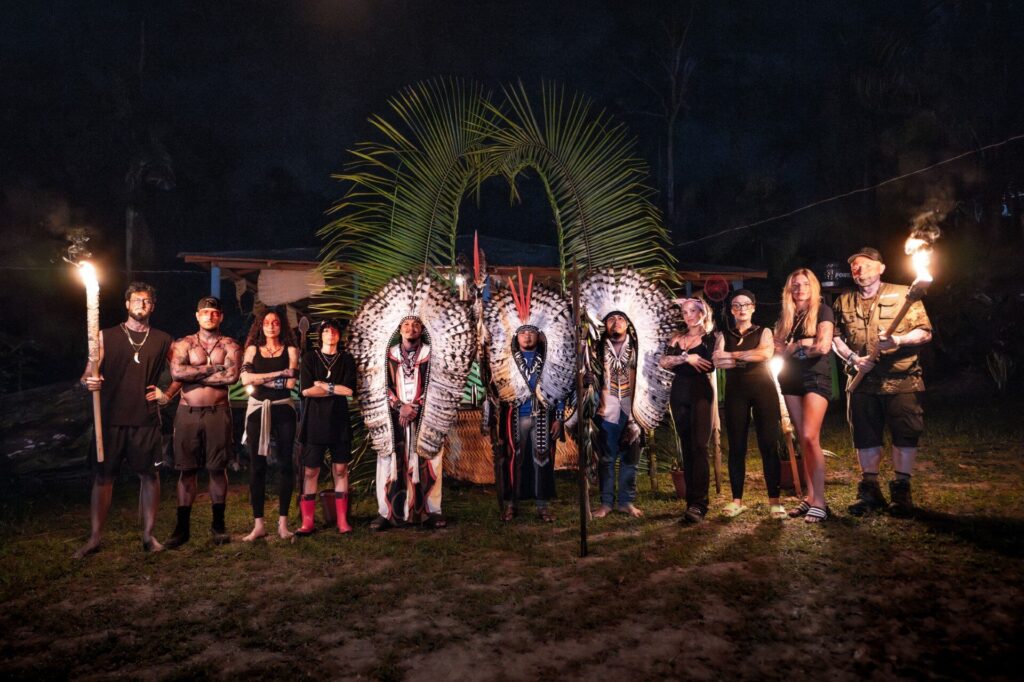 Gruppe von Menschen in traditioneller Kleidung mit Fackeln und Federschmuck in einem Waldgebiet.