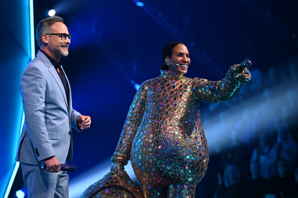 Zwei Personen auf einer Bühne, eine in einem glitzernden Kostüm und die andere in einem Anzug, während sie miteinander interagieren.