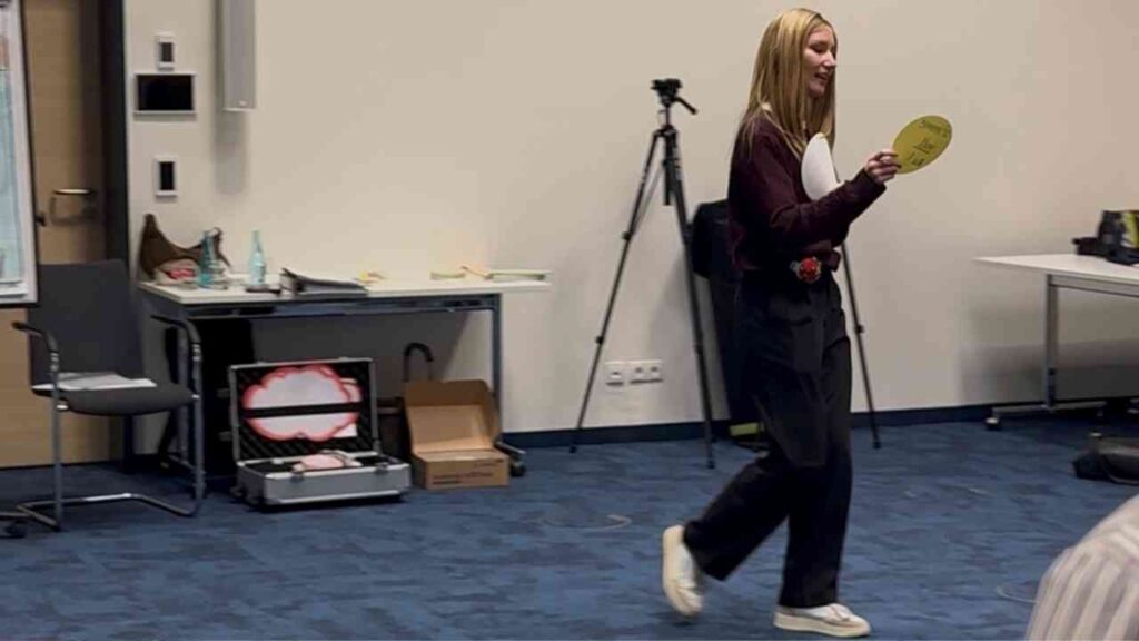 Junge Frau mit einem gelben Schild, die in einem Raum steht und spricht, während sie sich bewegt.