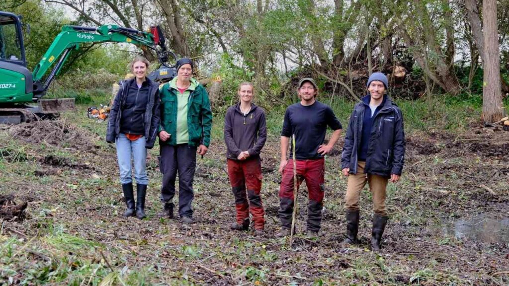Fünf Personen stehen in einer Landschaft mit einem Bagger im Hintergrund, während sie an einem Biotop arbeiten.
