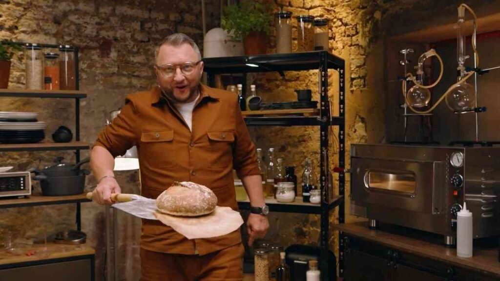 Ein Mann in einem braunen Outfit hält ein frisch gebackenes Brot auf einem Backblech in einer modernen Küche.