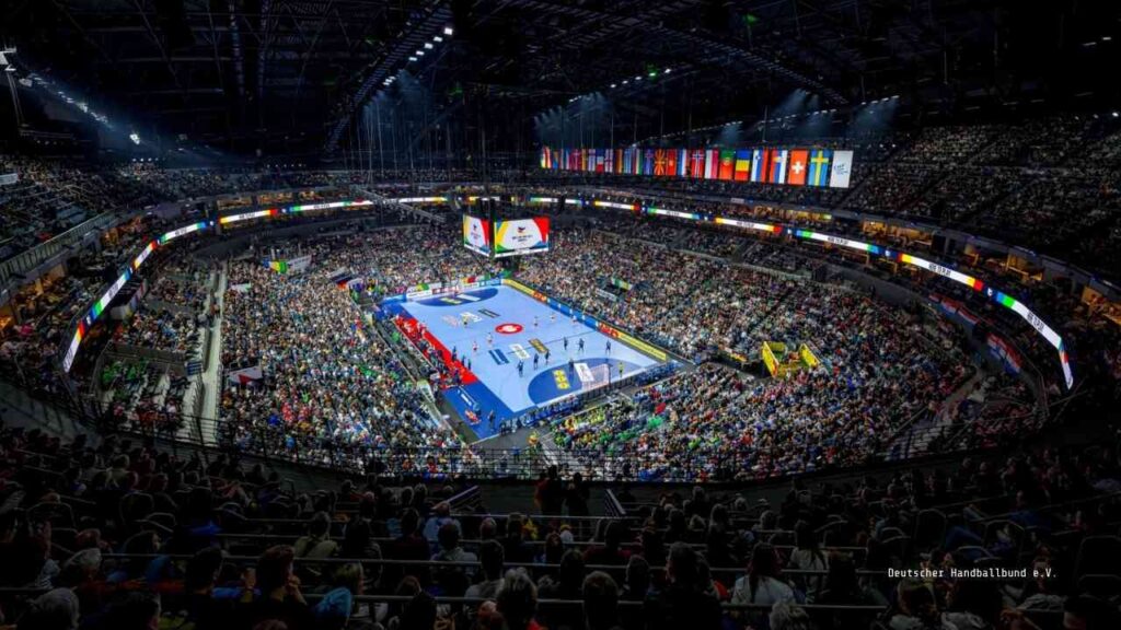 Voll besetzte Arena während eines Handballspiels mit Spielern auf dem Spielfeld und Zuschauern auf den Tribünen.