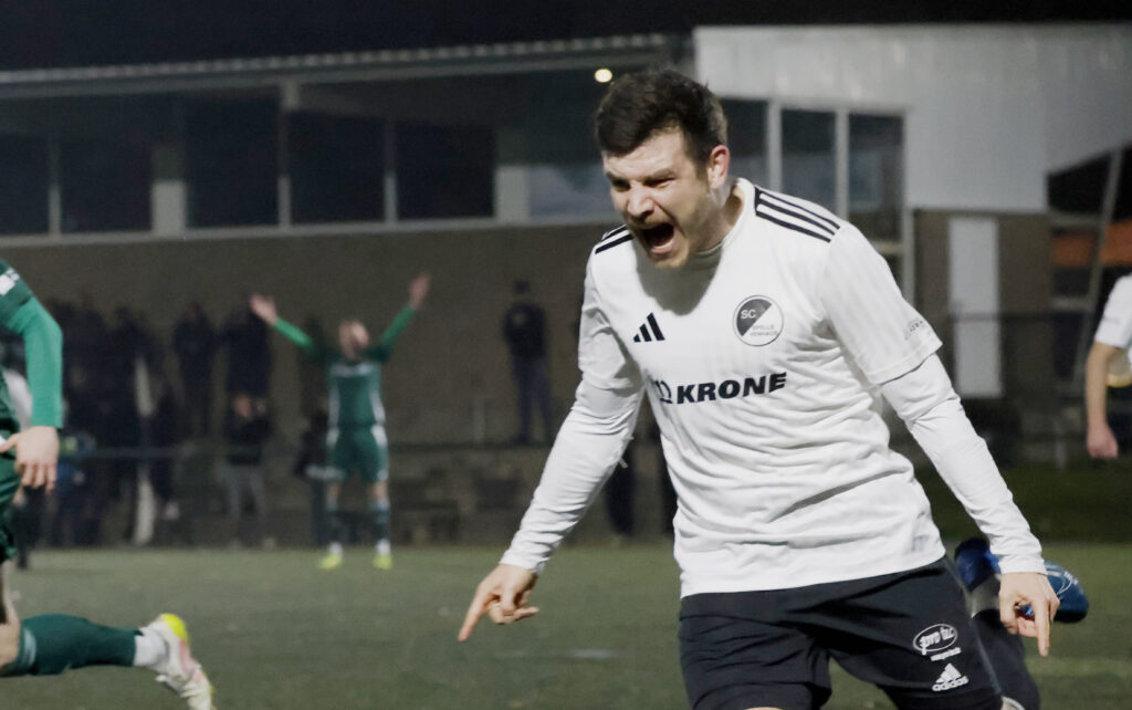 Fußballspieler in weißem Trikot, der nach einem Tor jubelt, während andere Spieler im Hintergrund agieren.