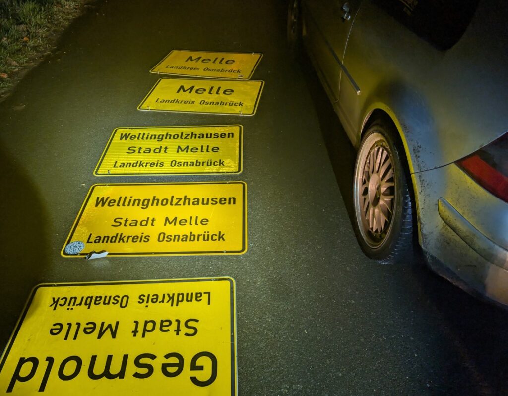 Gelbe Ortsschilder mit den Aufschriften 'Melle' und 'Wellingholzhausen' im Landkreis Osnabrück auf einer Straße.