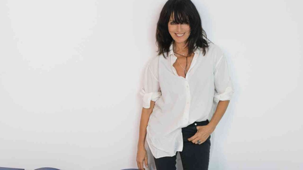 Frau mit langen dunklen Haaren trägt ein weißes Hemd und steht lächelnd an einer Wand.