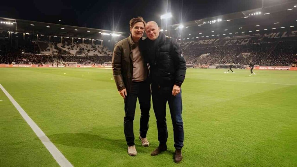 Zwei Männer stehen auf dem Rasen eines Fußballstadions vor einer Menschenmenge während eines Spiels.