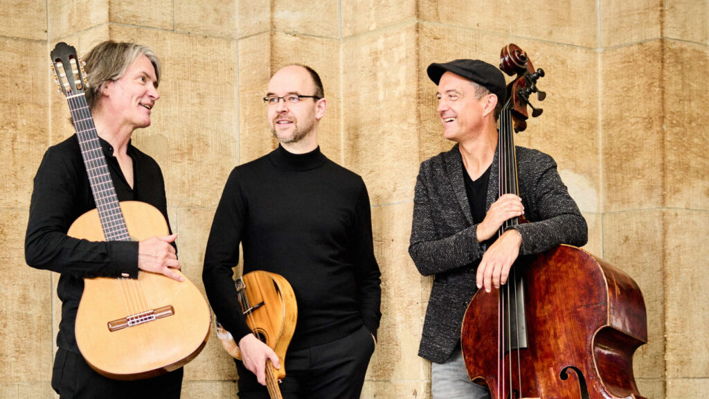 Drei Musiker stehen zusammen, einer mit einer Gitarre, einer mit einer Mandoline und einer mit einem Kontrabass.
