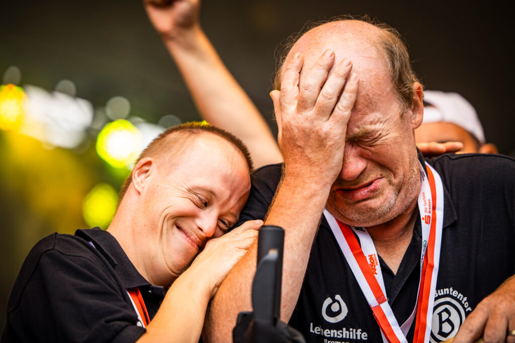 Zwei Männer mit Down-Syndrom, einer lächelt und der andere hält sich das Gesicht, beide tragen Medaillen.
