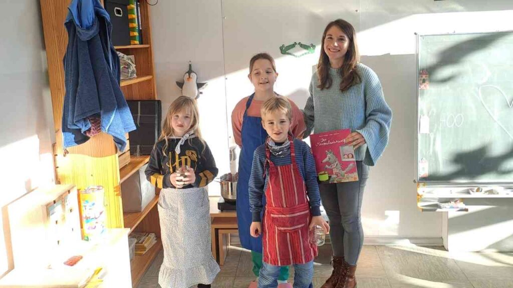 Vier Kinder und eine Erwachsene in einem Klassenzimmer, die ein Buch halten und in Schürzen gekleidet sind.