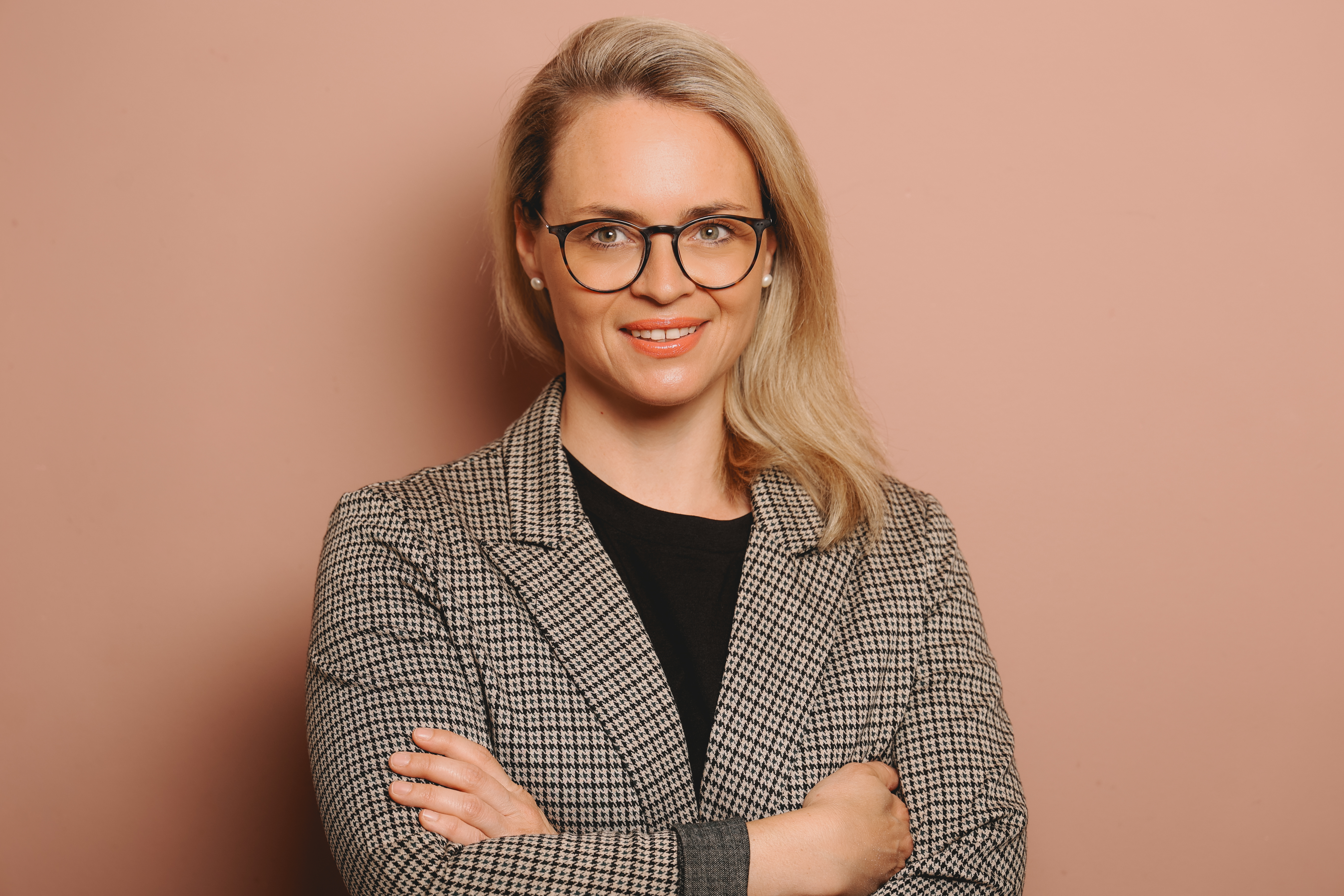 Porträt einer Frau mit blonden Haaren und Brille, die in einem karierten Blazer vor einem rosa Hintergrund steht.