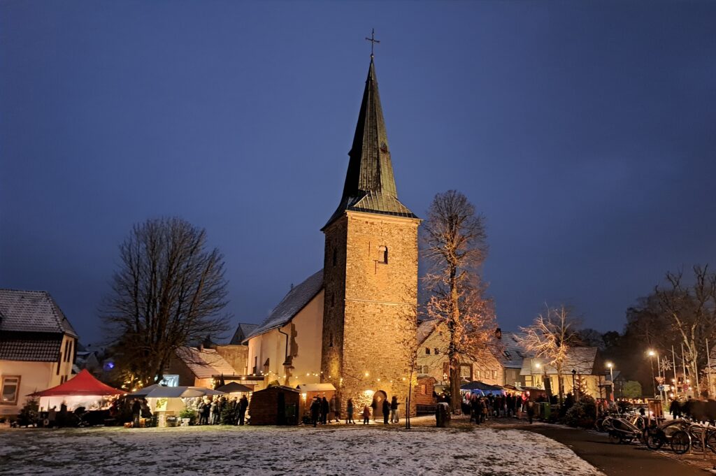 Kirchturm mit spitzem Dach und beleuchteten Fenstern in einer winterlichen Umgebung bei Nacht.