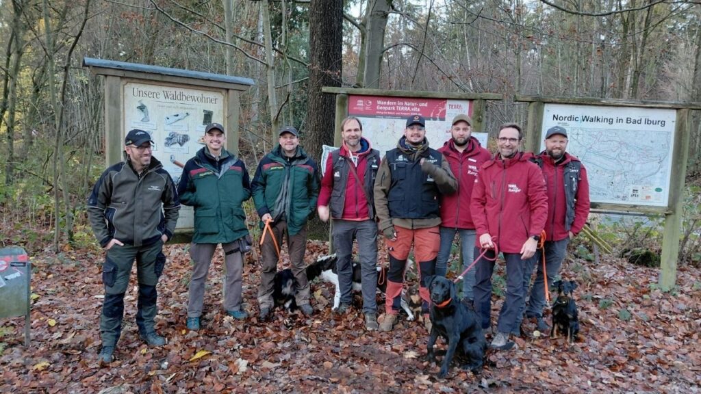 Gruppe von sieben Männern mit Hunden in einem Waldgebiet, gekleidet in Outdoor-Bekleidung, vor einem Informationsschild.