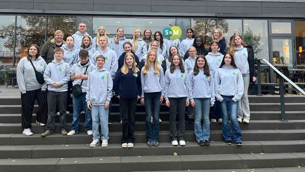 Gruppe von Jugendlichen und Erwachsenen in grauen Sweatshirts vor einem Gebäude mit dem Logo von SaM.