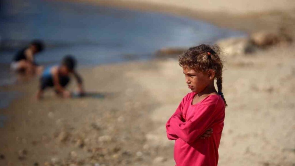 Ein Mädchen mit lockigem Haar und rotem Oberteil steht am Strand, während andere Kinder im Hintergrund spielen.