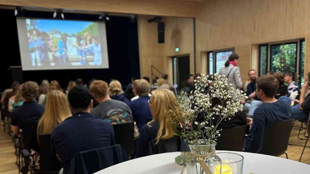 Gruppe von Menschen, die in einem Veranstaltungsraum auf einer Präsentation sitzen, mit einer Blumenvase im Vordergrund.
