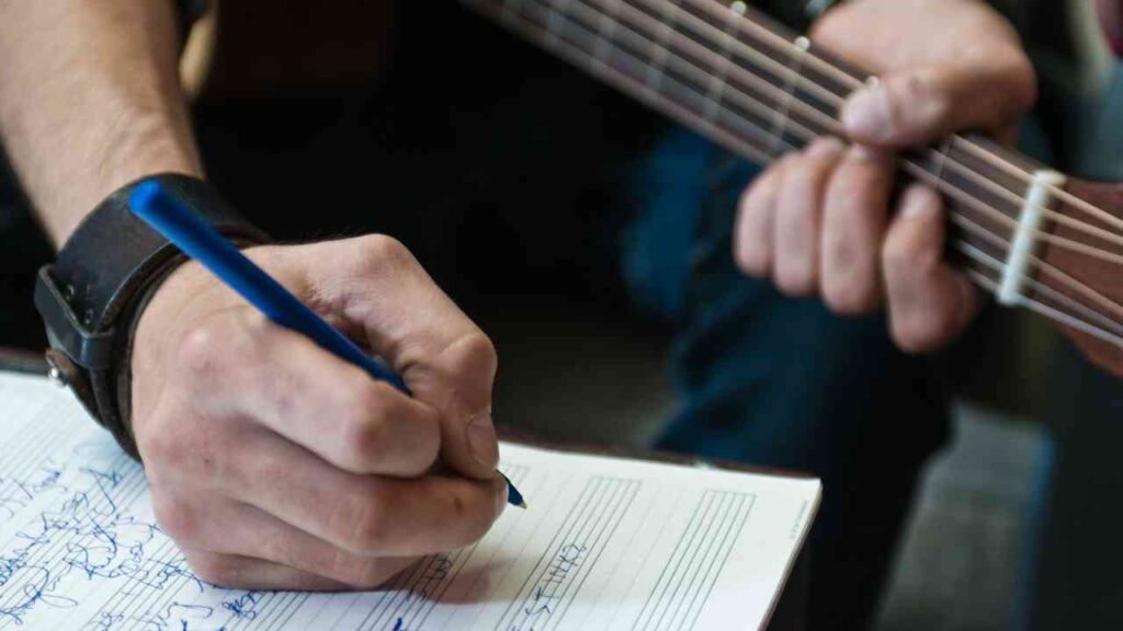 Händed eines Songwriters, der auf einem Notizblatt mit einem blauen Stift Notizen macht, während er eine Gitarre hält.