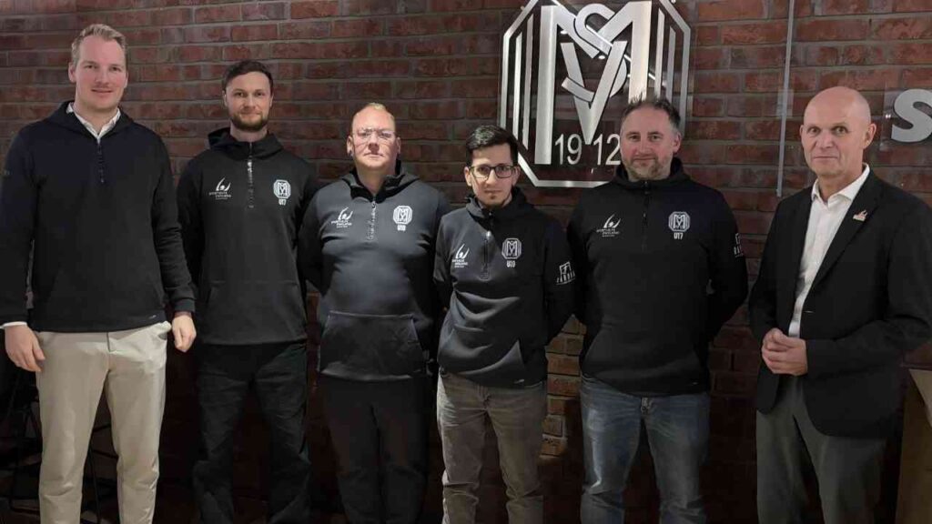 Gruppe von sechs Personen in schwarzen Kapuzenpullis, die vor einer Backsteinwand mit einem Logo stehen.