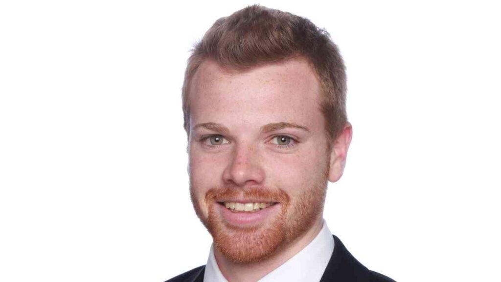 Porträt eines Mannes mit rotem Bart und kurzen, blonden Haaren in einem Anzug vor weißem Hintergrund.