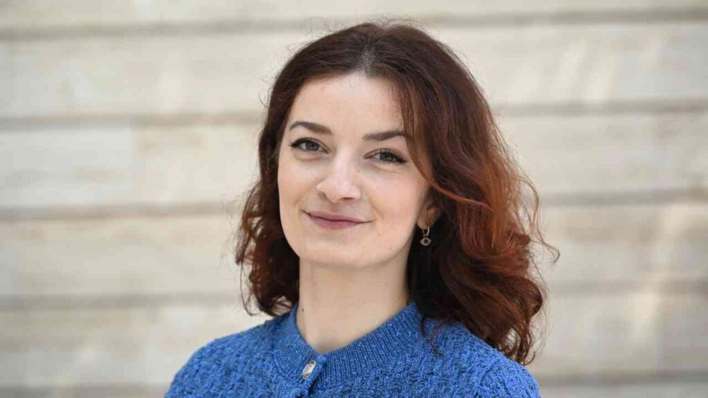 Frau mit lockigem, kastanienbraunem Haar und blauer Strickjacke, die in die Kamera lächelt.