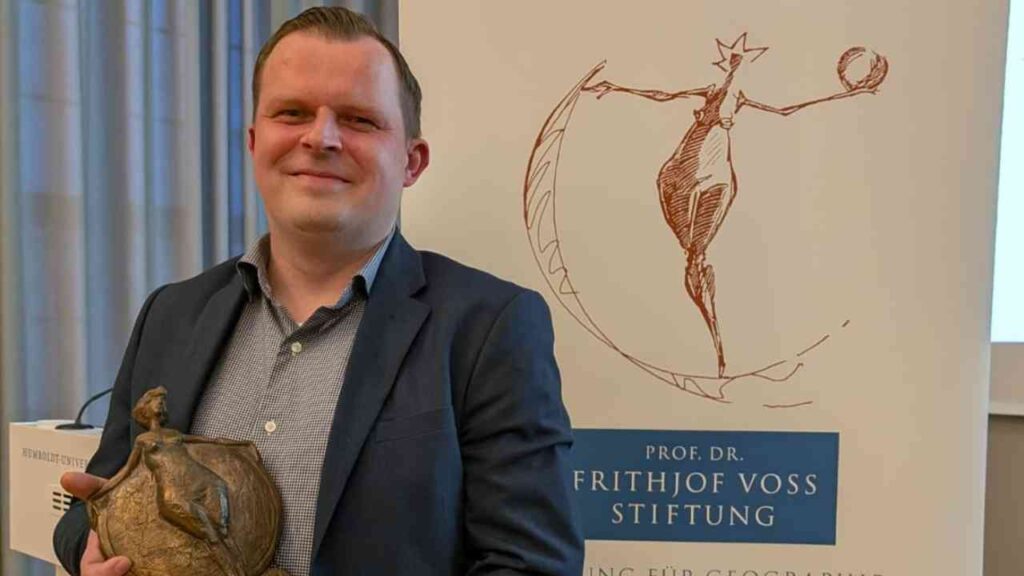 Mann in Anzug hält eine Auszeichnung der Frithjof Voss Stiftung in der Hand.