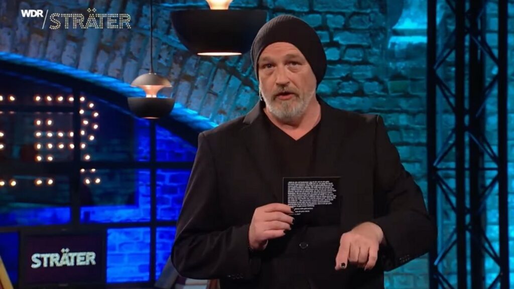 Mann mit schwarzer Mütze und Anzug hält eine Karte in der Hand und spricht auf einer Bühne mit Backsteinwand.