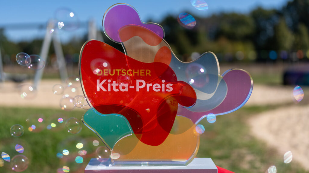 Transparente Auszeichnung in bunten Farben mit der Aufschrift 'Deutscher Kita-Preis' auf einem Tisch im Freien.