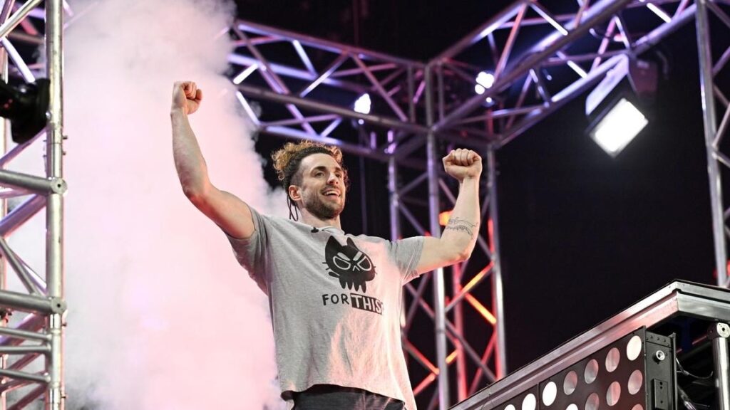 Mann mit hochgestreckten Armen auf einer Bühne, umgeben von Nebel und Lichtern, trägt ein graues T-Shirt mit Aufdruck.