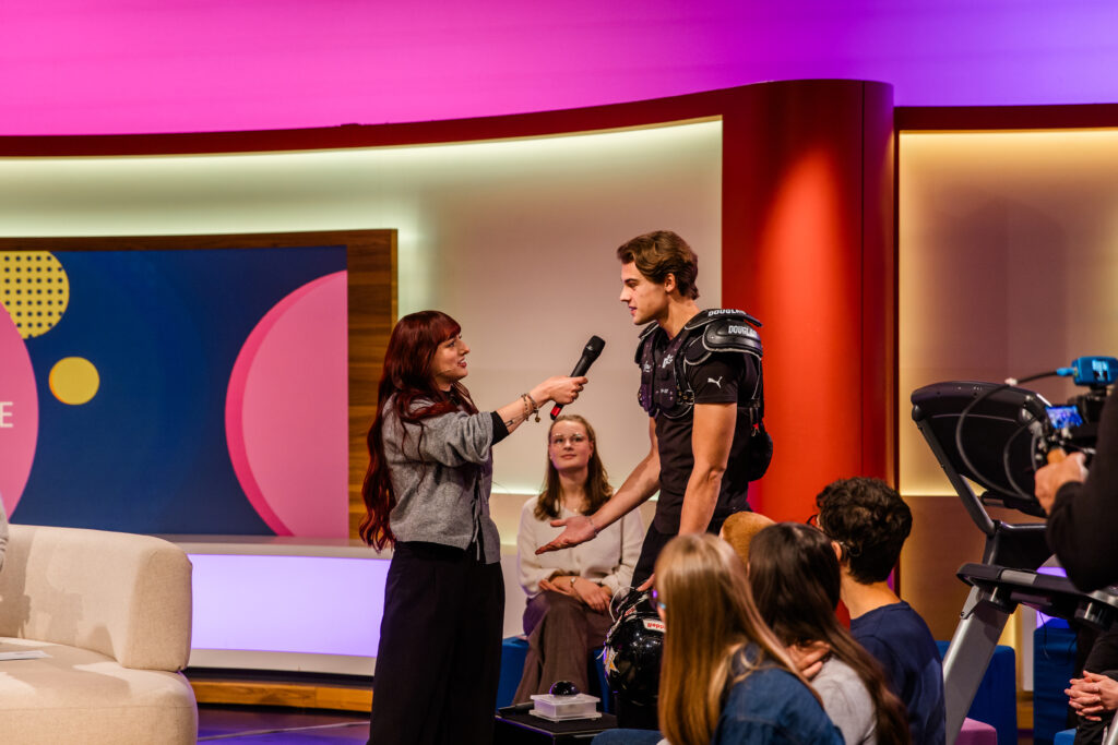 Eine Frau interviewt einen Mann in einem futuristischen Anzug während einer Fernsehsendung.