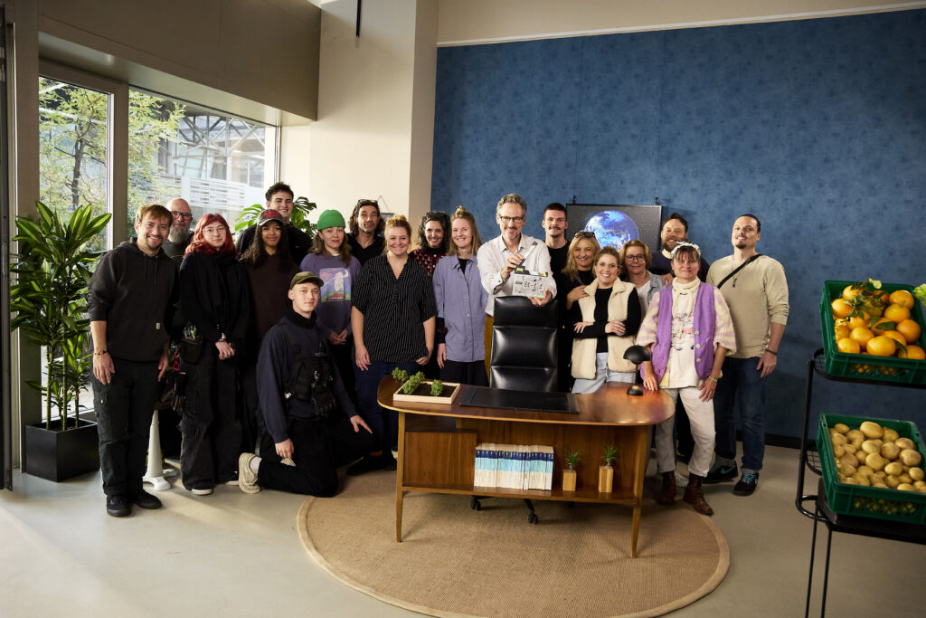 Gruppe von Personen, die um einen Tisch in einem modernen Raum versammelt sind, mit verschiedenen Pflanzen und Obstkörben im Hintergrund.