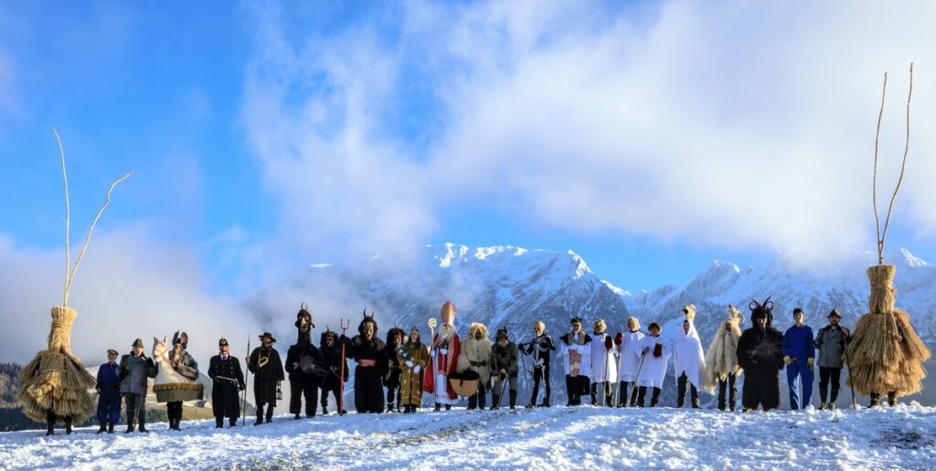 Gruppe von Menschen in traditionellen Kostümen, die auf einem schneebedeckten Hang stehen, mit Bergen im Hintergrund.