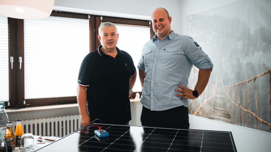 Zwei Männer stehen neben einem Solarpanel in einem Raum mit Fenstern und natürlichem Licht.