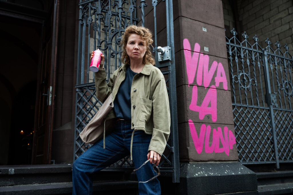 Frau mit lockigem Haar in einer beigen Jacke, die ein Getränk hält, vor einer Wand mit pinkem Graffiti 'VIVA LA VULP'.