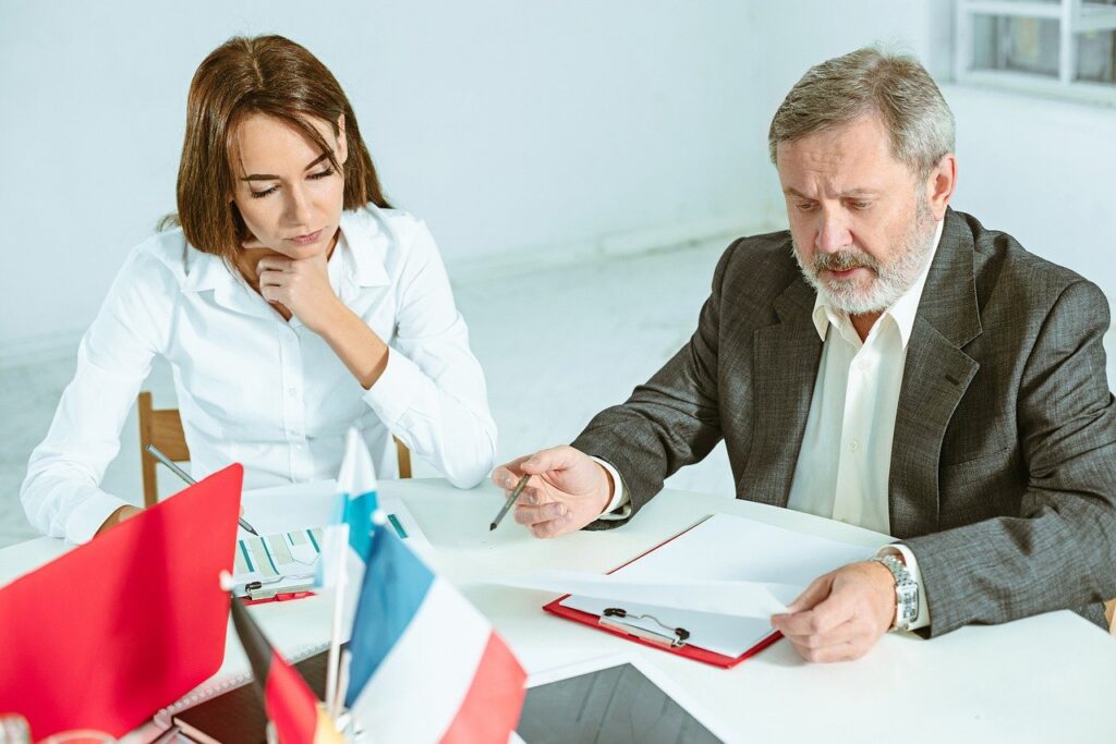 Zwei Personen in einem Besprechungsraum, die Dokumente und Notizen auf einem Tisch überprüfen, mit Flaggen von Deutschland und Frankreich im Hintergrund.