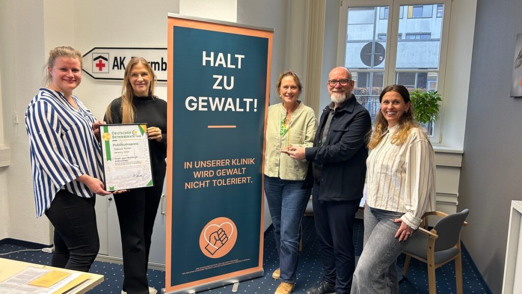 Gruppe von fünf Personen vor einem Banner mit der Aufschrift 'Halt zu Gewalt!' in einer Klinik.