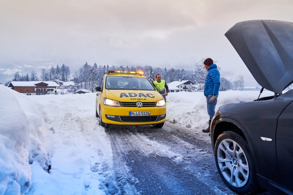 Gelbes ADAC-Fahrzeug mit Warnlichtern auf einer verschneiten Straße in den Alpen, während ein Mann mit einem anderen Auto spricht.