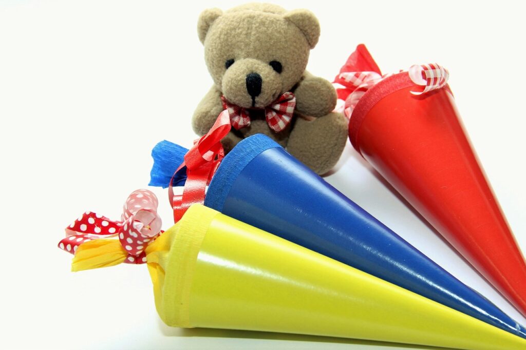 Drei bunte Schultüten in Gelb, Blau und Rot mit einem Teddy in der Mitte, der eine Schleife trägt.