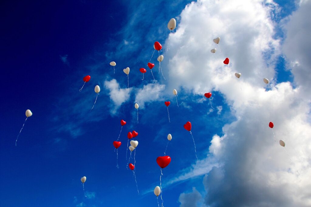 Rote und weiße Herzballons, die in den blauen Himmel mit Wolken aufsteigen.