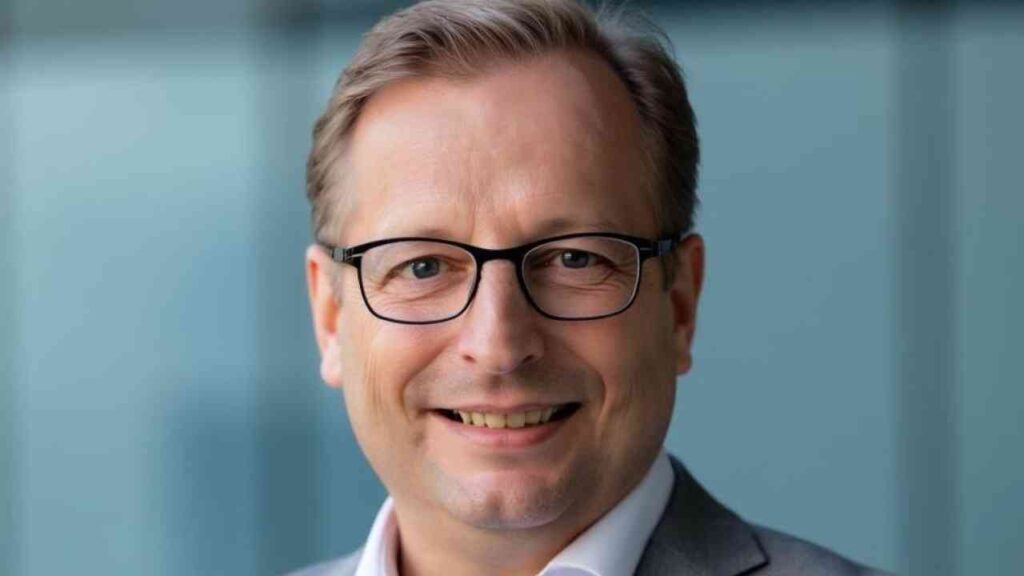 Mann mit Brille und Anzug, lächelnd vor einem unscharfen Hintergrund.