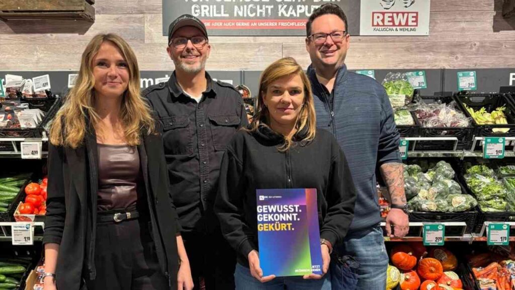 Vier Personen stehen in einem Supermarkt vor einem Regal mit frischem Gemüse und halten ein Buch mit dem Titel 'Gewusst. Gekonnt. Gekürt.'