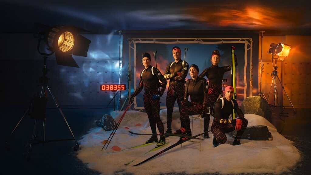 Fünf Biathleten in sportlicher Kleidung mit Skiern und Stöcken in einem Studio-Setting.