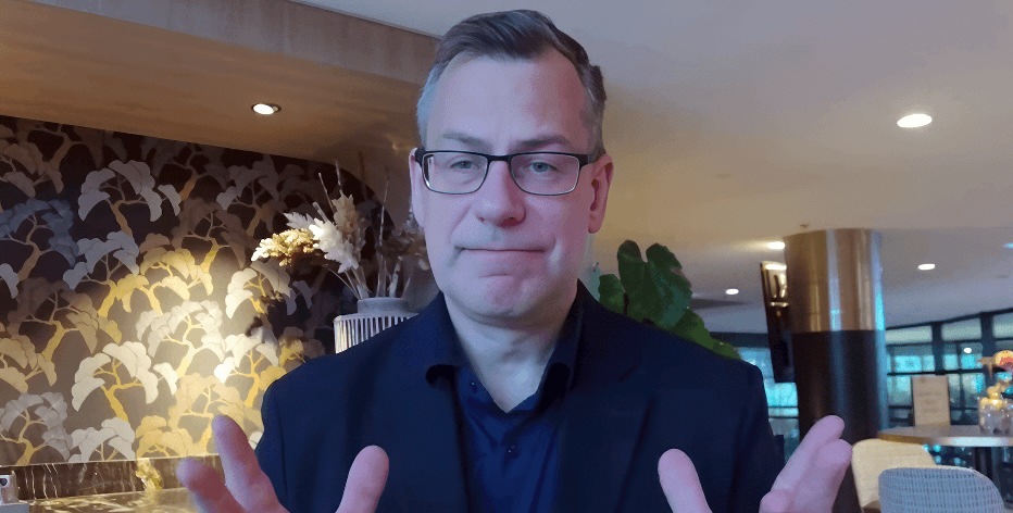 Mann mit Brille in einem modernen Innenraum, der mit beiden Händen gestikuliert.