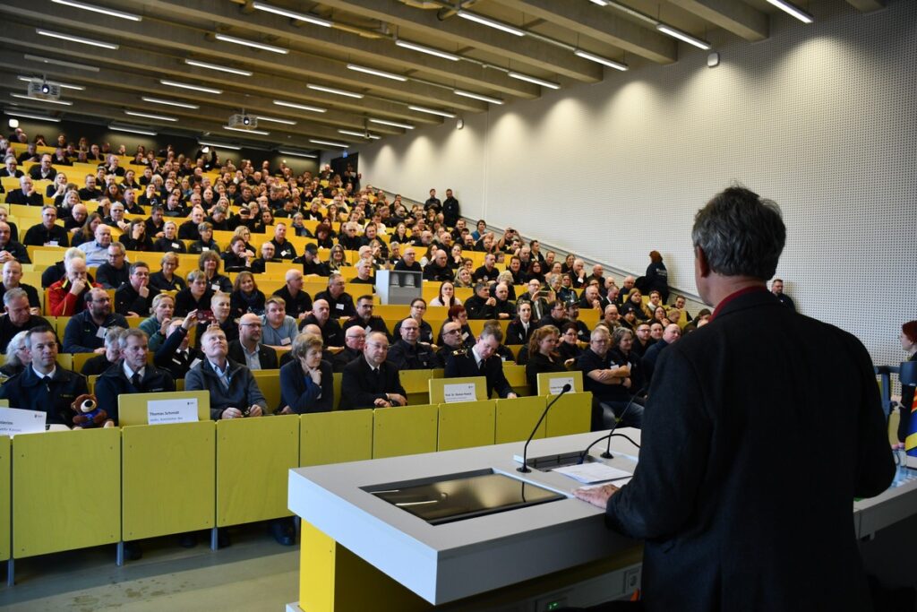 Ein Redner steht vor einem großen Publikum in einem modernen Hörsaal mit gelben Sitzplätzen.