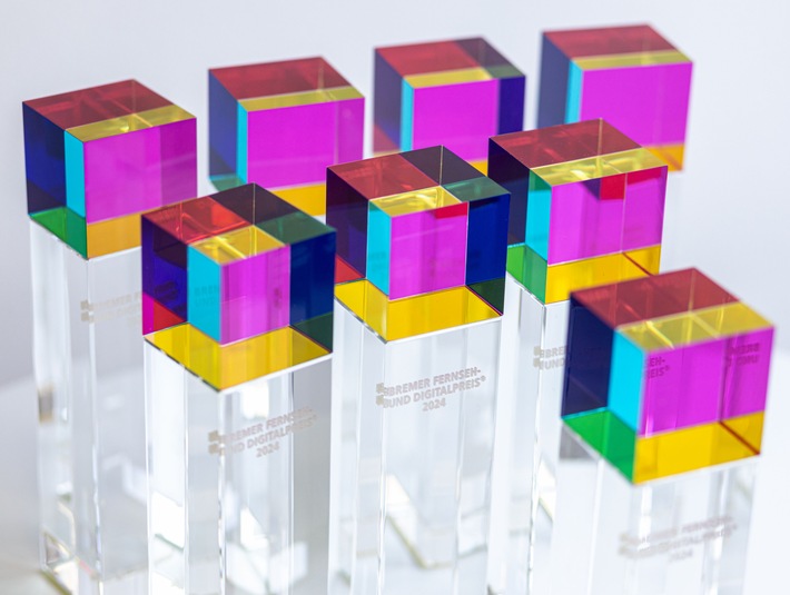 Acht farbige Auszeichnungen in Form von klaren Stelen mit bunten, quadratischen Oberteilen in Lila, Gelb und Grün.