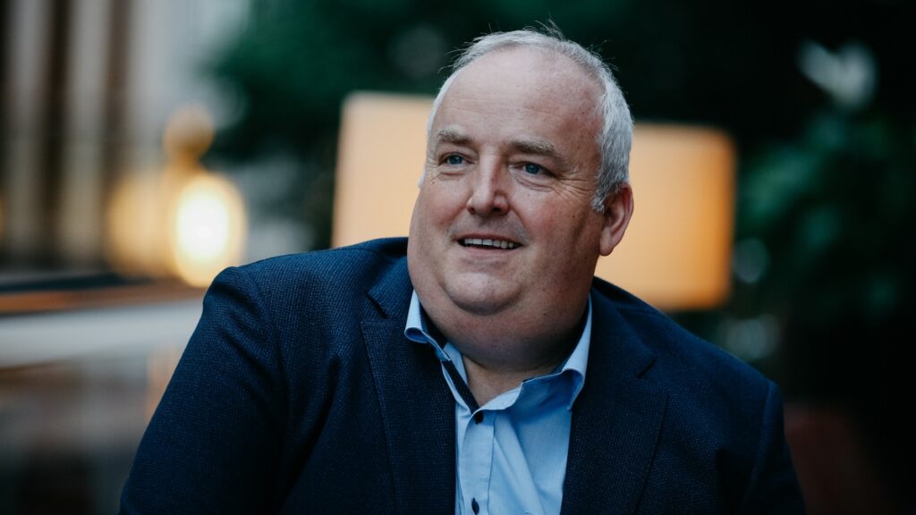 Mann mit kurzen grauen Haaren und einem blauen Anzug, der in einem Innenraum lächelt.