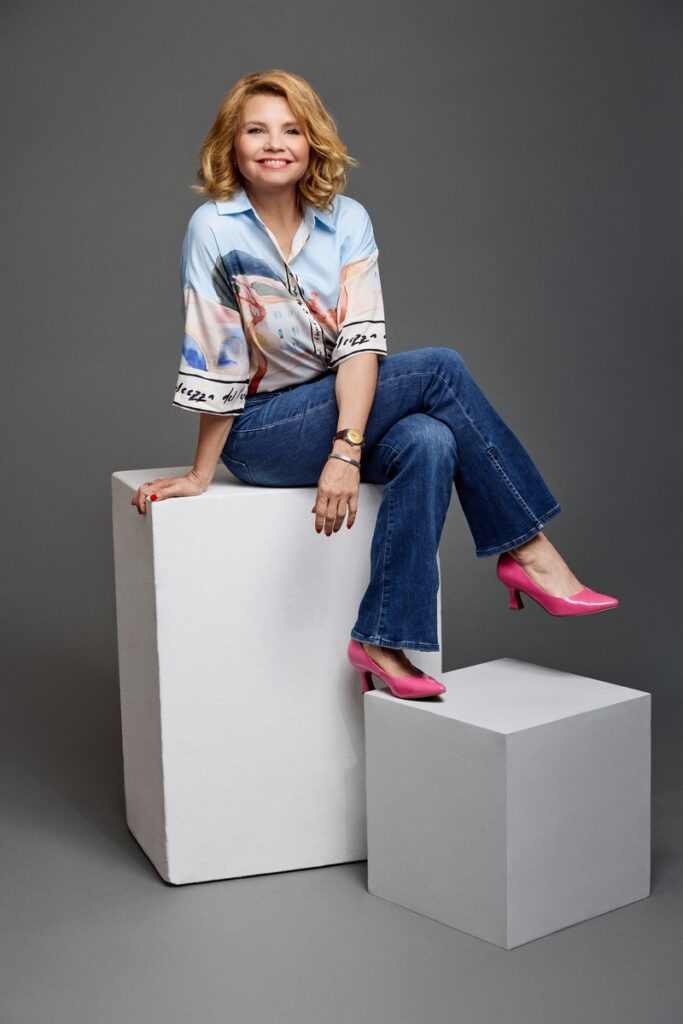 Frau mit blonden Locken in einem bunten Hemd, lächelnd und in einer entspannten Pose vor einem grauen Hintergrund.