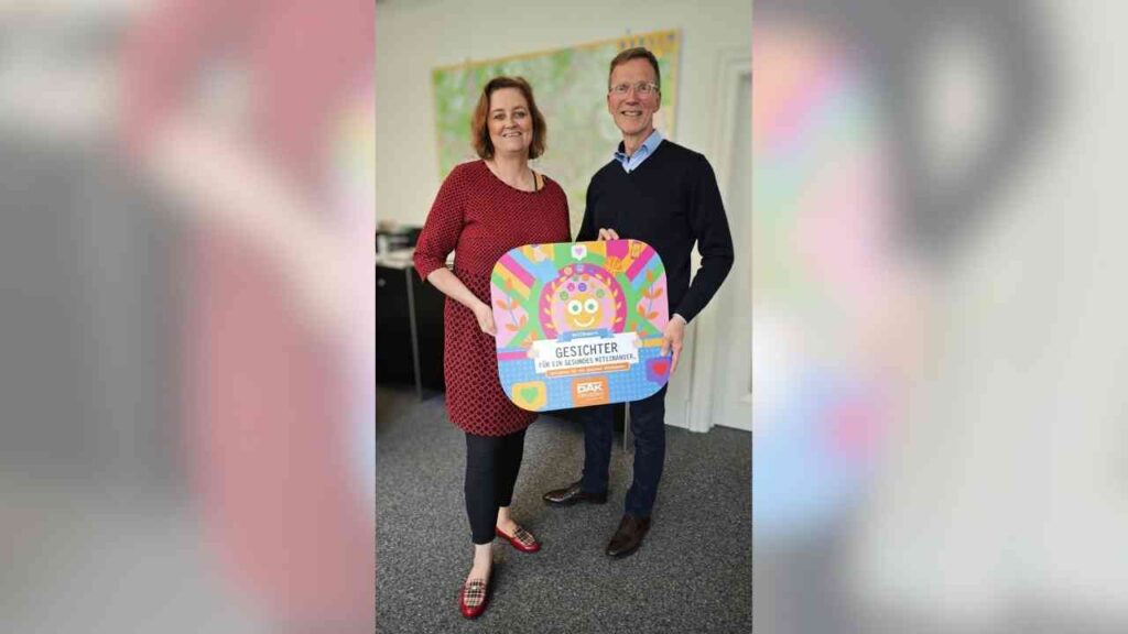 Zwei Personen halten ein buntes Schild mit der Aufschrift 'Gesichter der Gemeinschaft' in einem Büro.