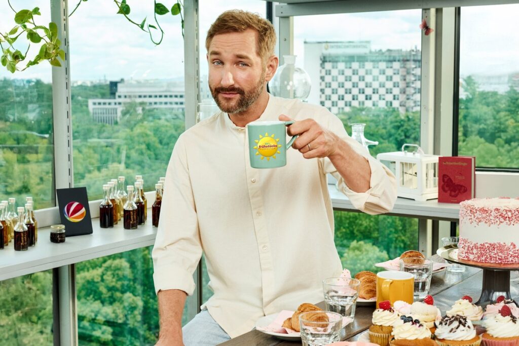 Mann mit Bart hält eine Tasse mit Sonnenmotiv in einer hellen Umgebung, umgeben von verschiedenen Speisen und Getränken.